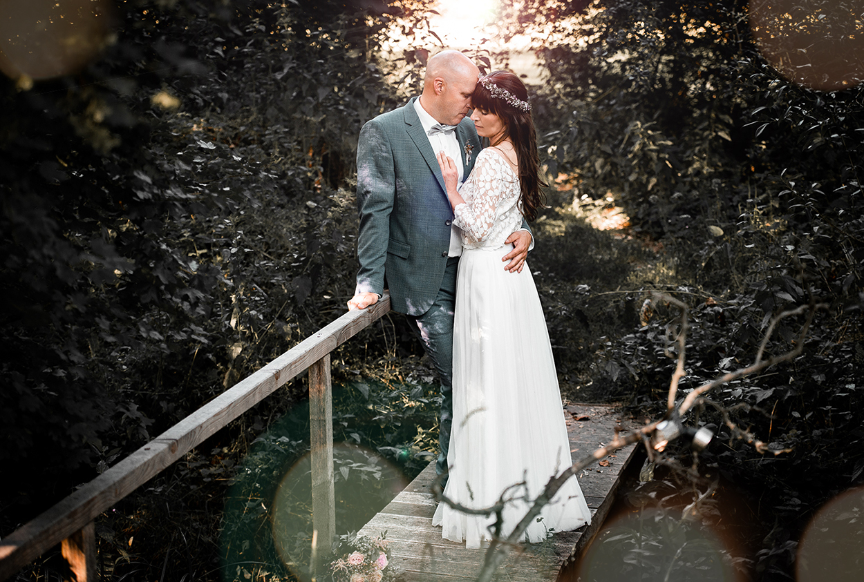 Brautpaar steht eng umarmt auf einer Holzbrücke im Grünen bei Sonnenlicht – natürliche Hochzeitsfotografie von Stefanie Cypris, Fotografin Nürnberg Schwabach Roth Fürth